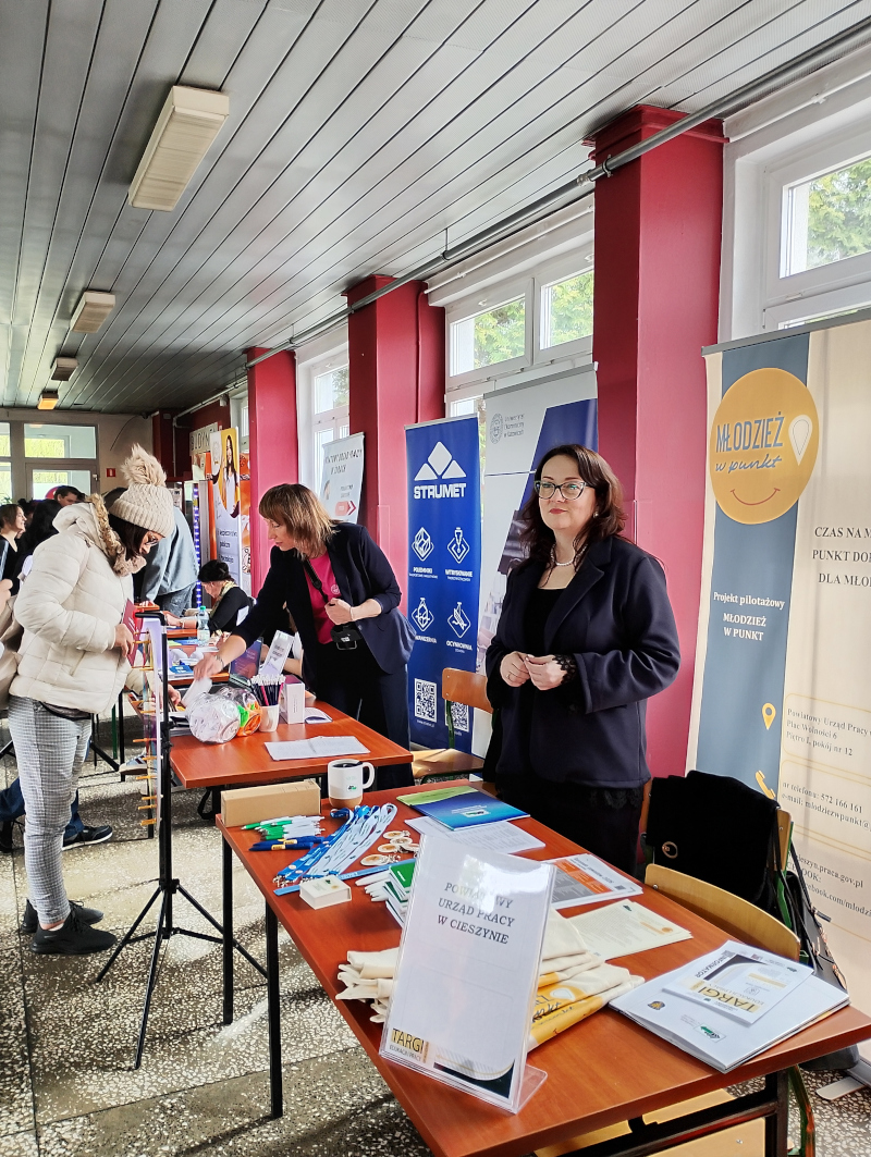 2.	Stoisko informacyjne podczas targów edukacji i pracy, na stolikach materiały promocyjne: ulotki, długopisy, smycze i broszury. Na pierwszym planie pracownica cieszyńskiego PUP za nią baner Młodzież w Punkt, w oddali inne stoiska i osoby odwiedzające targi.