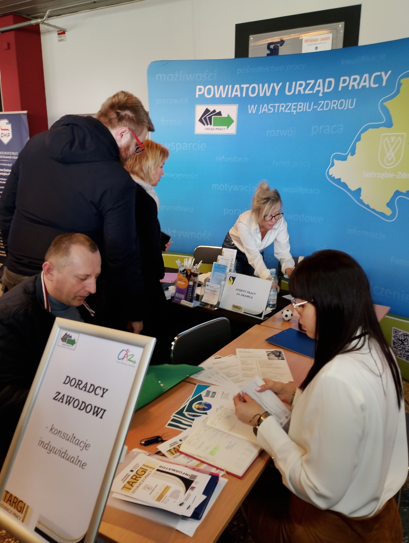 3.	Przy stoisku doradcy zawodowi z PUP w Jastrzębiu Zdroju wraz z osobami odwiedzającymi punkt konsultacyjny na targach edukacji i pracy, w tle baner urzędu wraz z logotypami.