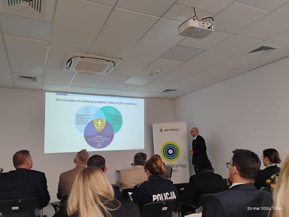 Grupa osób uczestnicząca w spotkaniu informacyjnym, patrząca na wyświetlany slajd dotyczący grupy docelowej projektu.