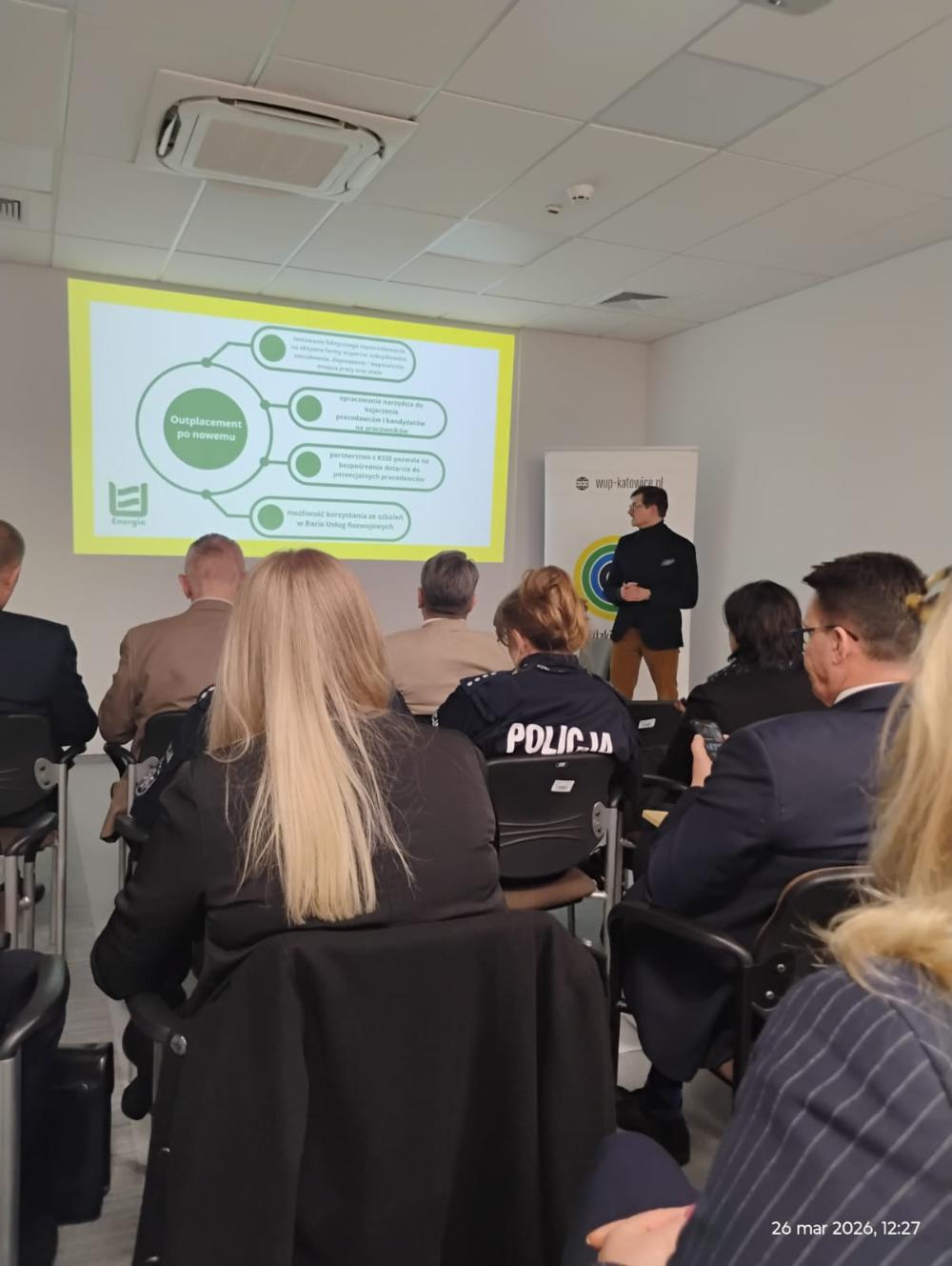 Grupa osób uczestnicząca w spotkaniu informacyjnym. Uczestnicy patrzą na slajd przedstawiający Outplacement po nowemu.